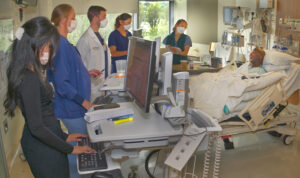 A patient lies in a hospital bed with a team of masked doctors standing nearby. One is typing on a large monitor in the foreground.