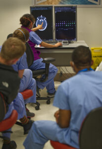 Small classroom of doctors facing toward the back of the room and observing a monitor, as a person close to screen points to an image.
