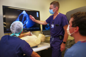 Three doctors observe a simulation with a mannequin, using a monitor.