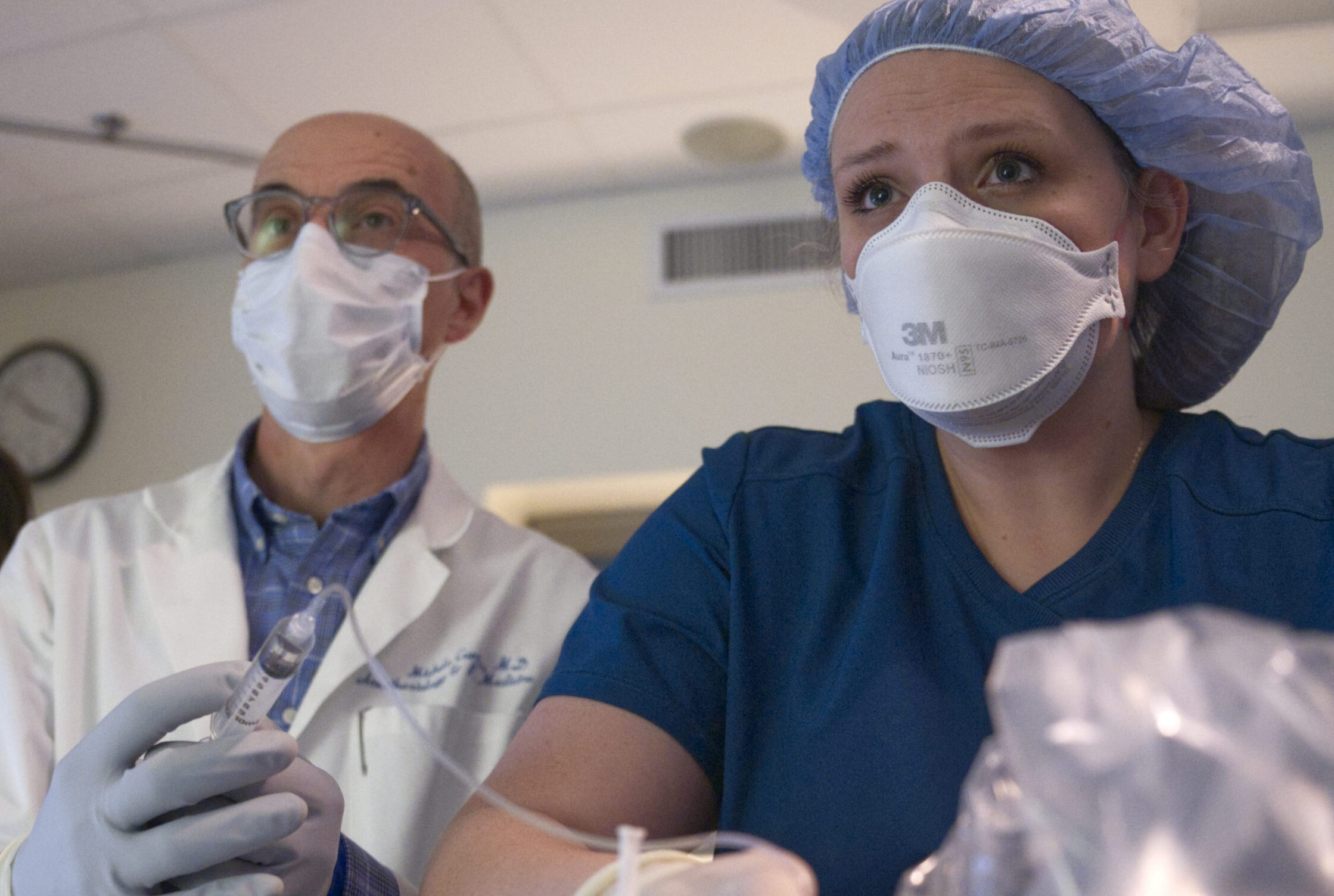 Male in white coat and female in blue scrubs and blue cap using a clear syringe.