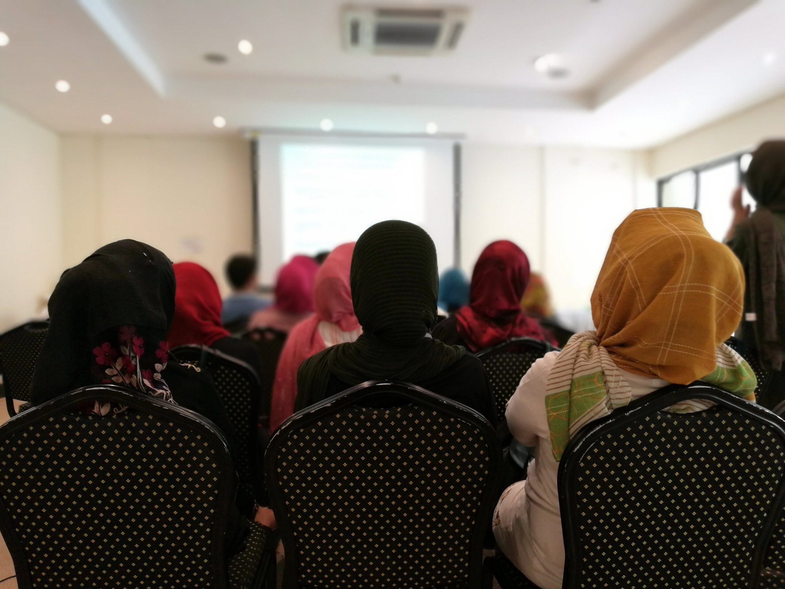 Three seated women wearing hijabs facing away from the camera watching a presentation at the front of the room.