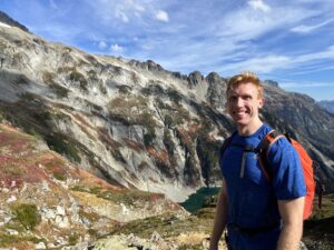 young man wearing blue shirt and orange backpack standing by mountain hiking trail 