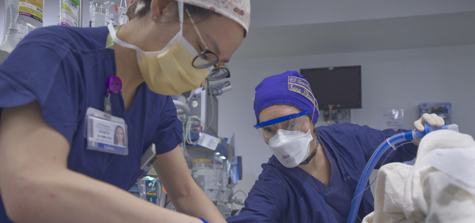 A trainee receiving instruction from Dr. Sara Nikravan. They are with an unseen patient and in protective outerwear.