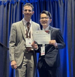Dr. Mukai dressed in a suit and standing on a podium, receiving his SNACC award certificate with the presenter.