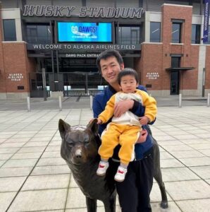 Yajun Wang, PhD, standing outside of Husky Stadium. He is holding a child wearing a yellow outfit.