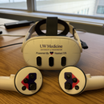 A headset and two hand controls imprinted with the UW Medicine and Vantari VR logos sits on a table by a window.