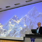 Dr. Mike Grocott standing at a lecturn. The large screen behind him projects a photo of Mt. Everest.