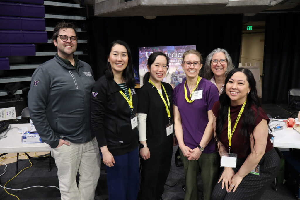 Six smiling volunteers and staff pose together at a backstage event, wearing name badges and yellow lanyards.