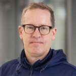 Portrait of a man with short light brown hair and glasses, wearing a navy zip-up jacket, looking at the camera.
