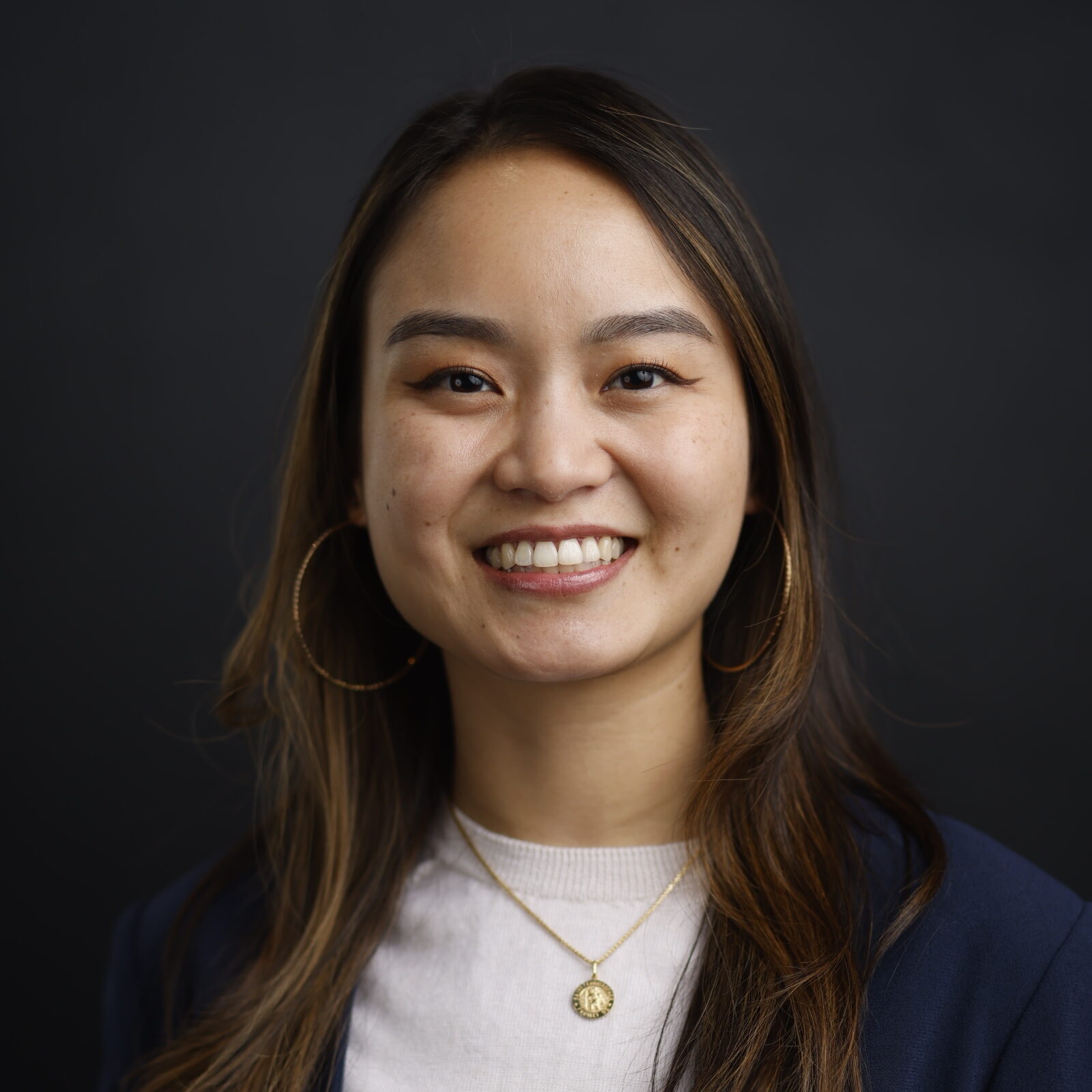 Portrait of a smiling woman with long hair, wearing a white top and dark blazer, against a dark background, gold hoop earrings visible.