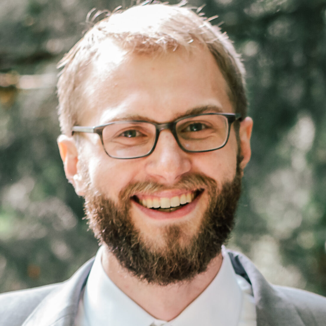 Smiling man with glasses and beard outdoors, blurred green background.