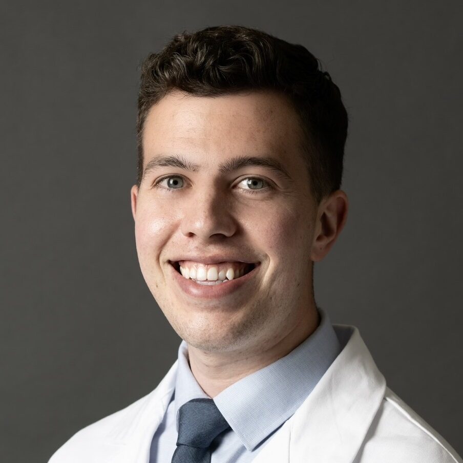 Smiling male doctor in a white coat and blue tie, posing against a dark gray studio backdrop (head-and-shoulders portrait).