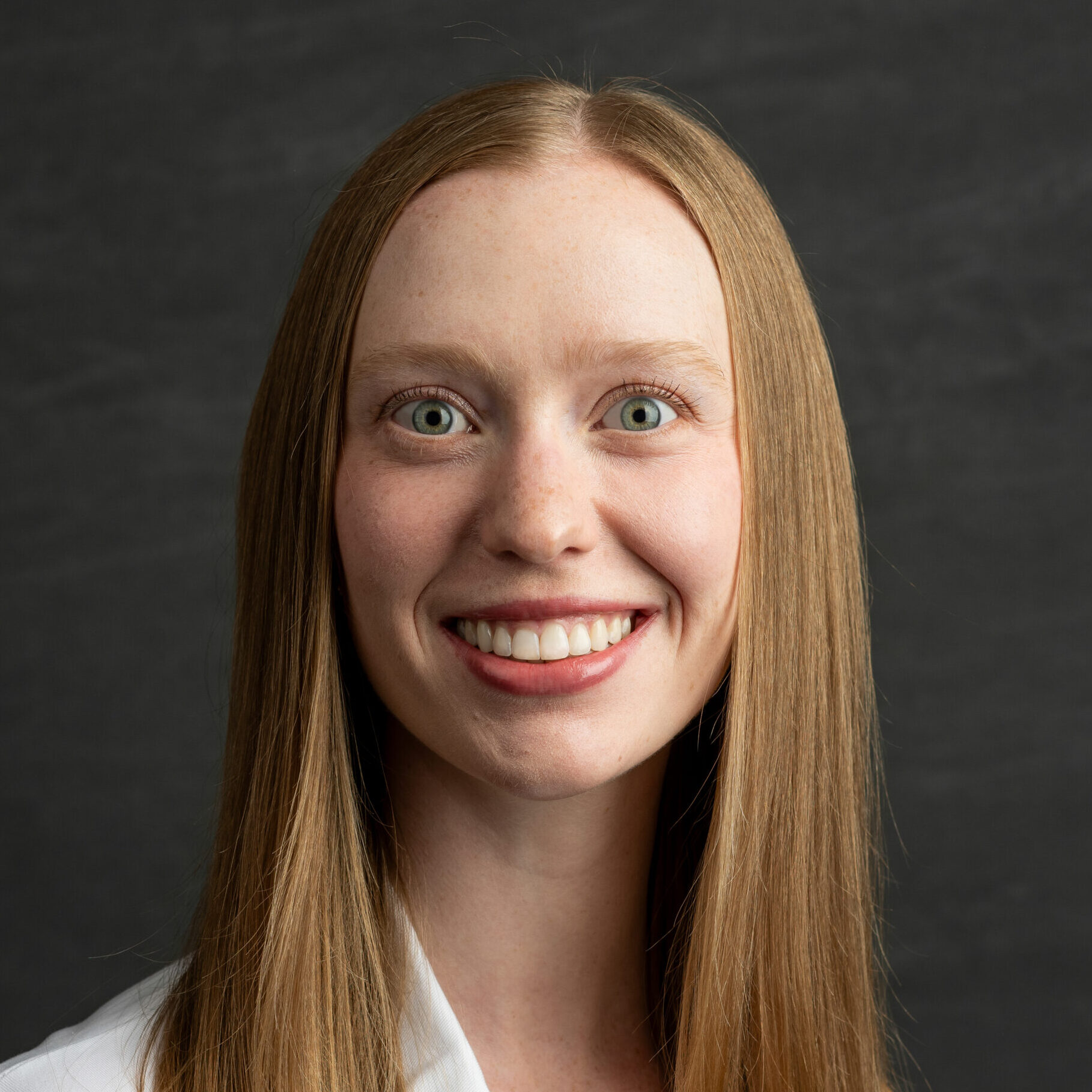 Close-up portrait of a smiling woman with long light brown hair wearing a white blazer against a dark gray background