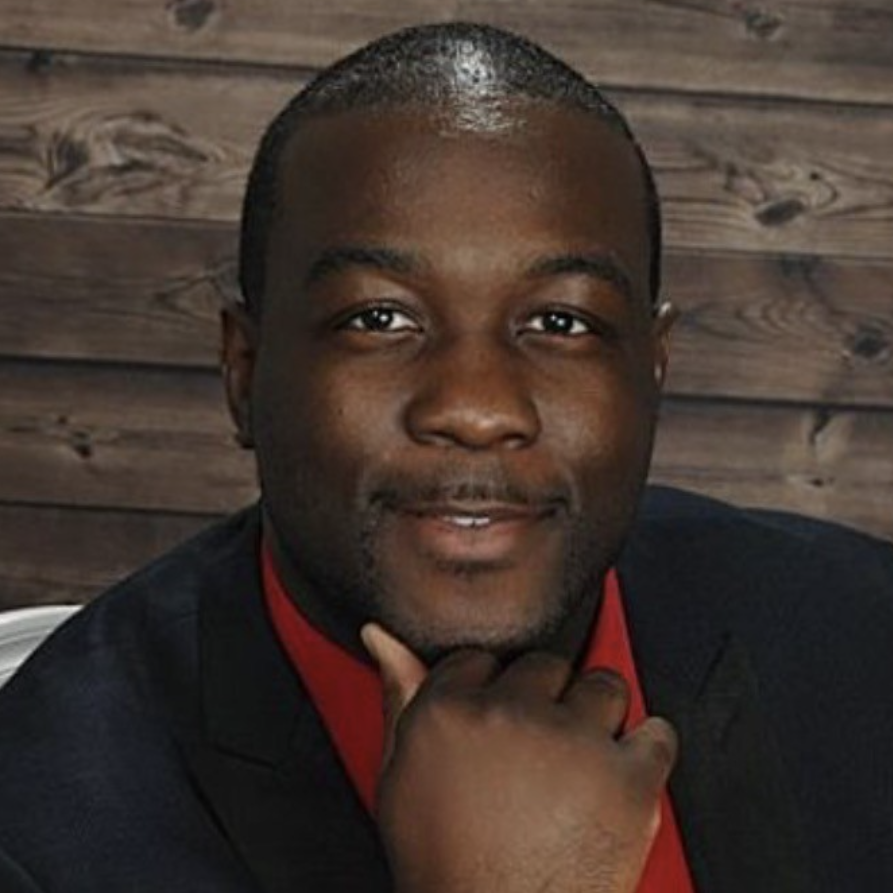 Head-and-shoulders portrait of a smiling Black man in a dark suit and red shirt, resting his chin on his hand against a wooden backdrop.