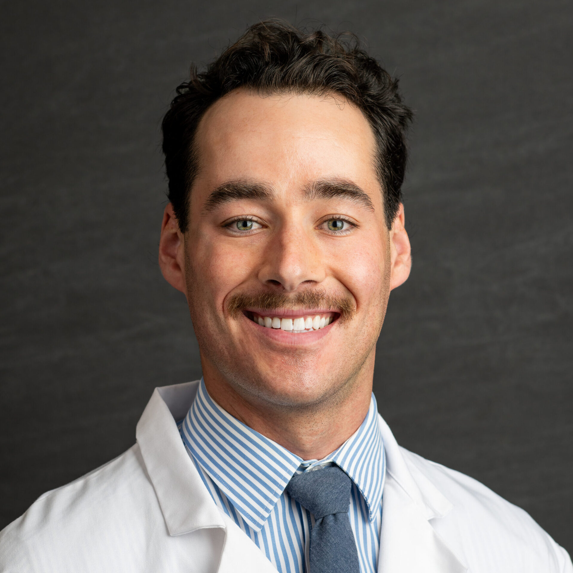 Smiling man in a white lab coat and blue striped shirt, Anesthesiology & Pain Medicine embroidery visible on pocket area.