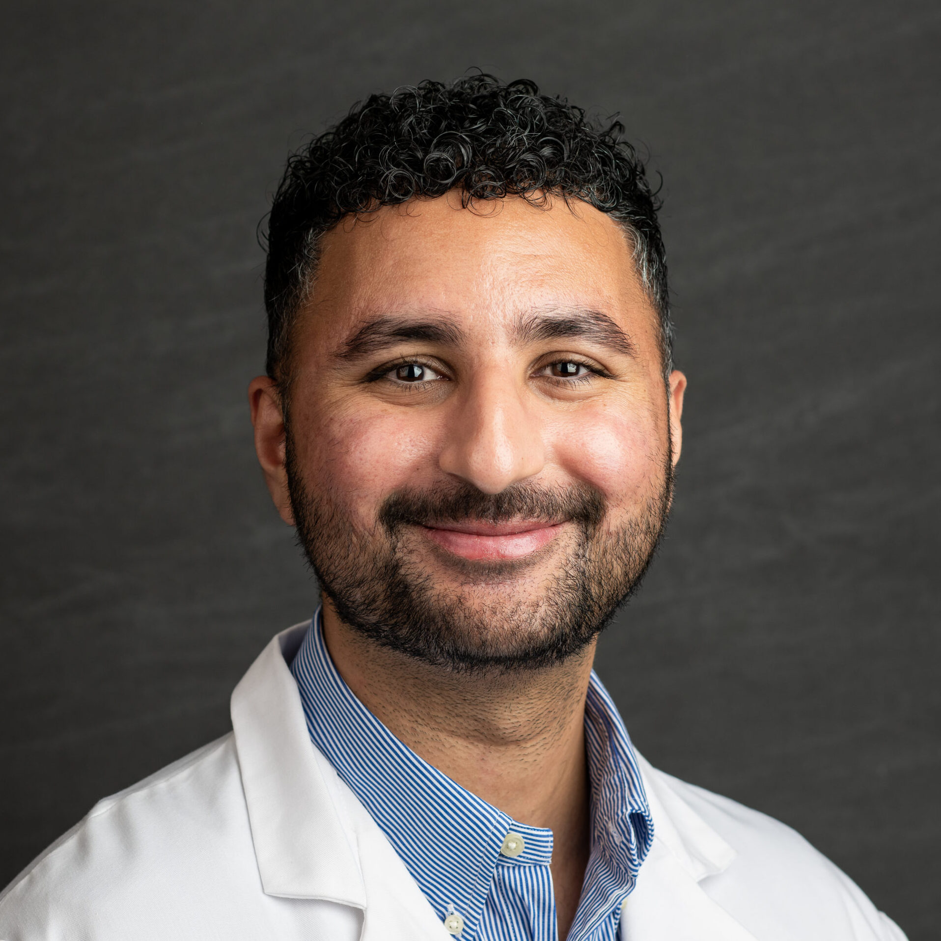 Smiling man in a white lab coat and blue striped shirt against a dark gray background (professional portrait).