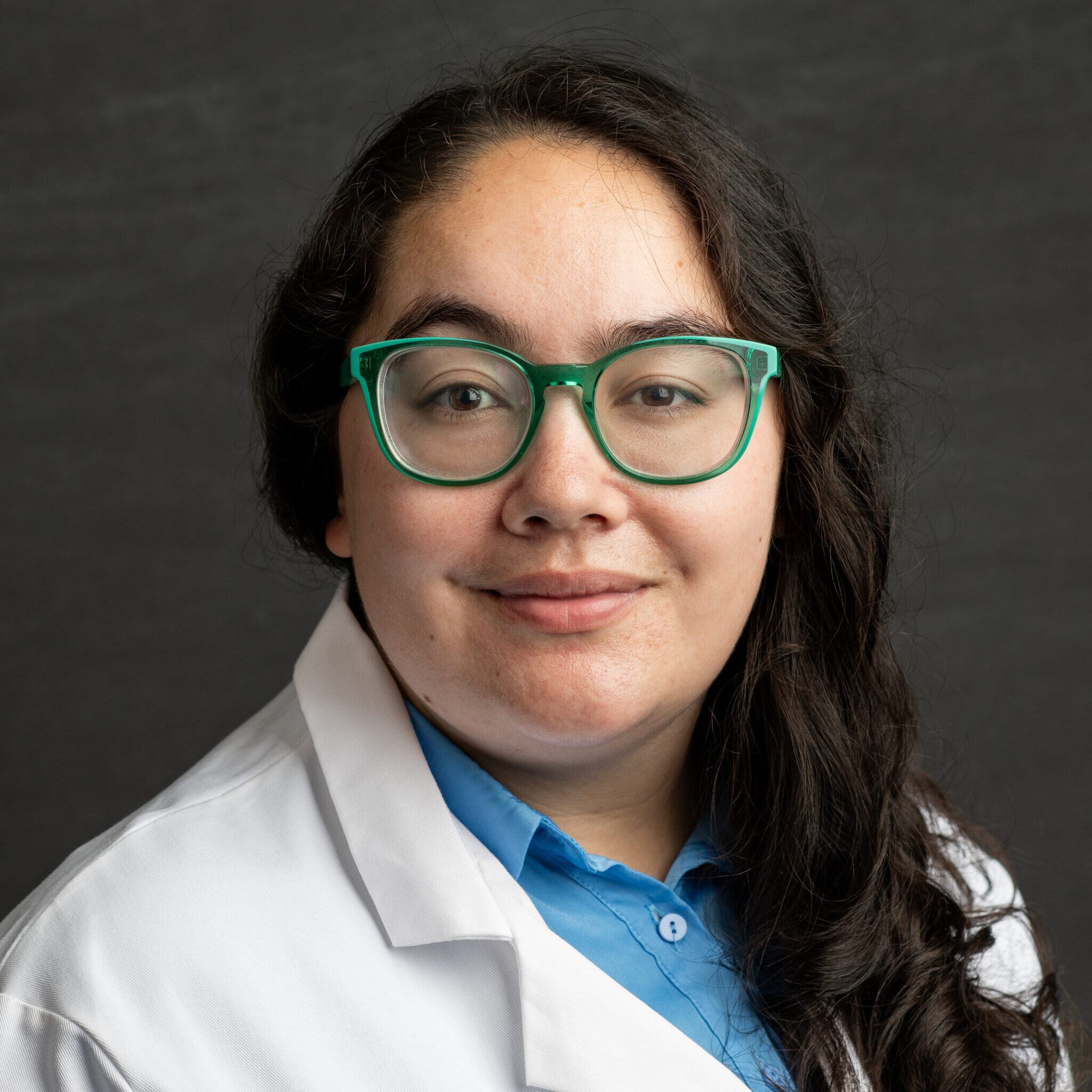 Person with long dark curly hair in doctor's coat wearing green glasses