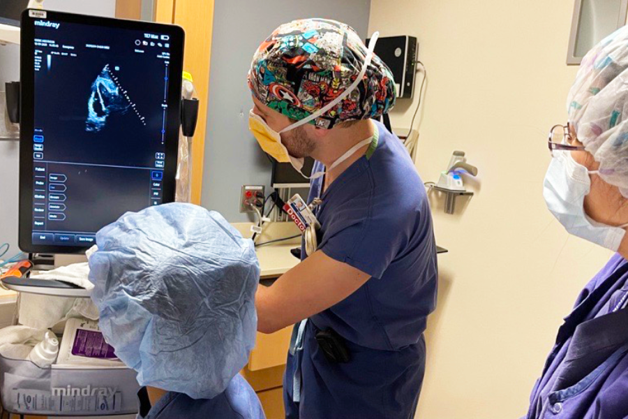 Two healthcare professionals in scrubs and masks operate an ultrasound machine in a clinical setting, reviewing the monitor display.