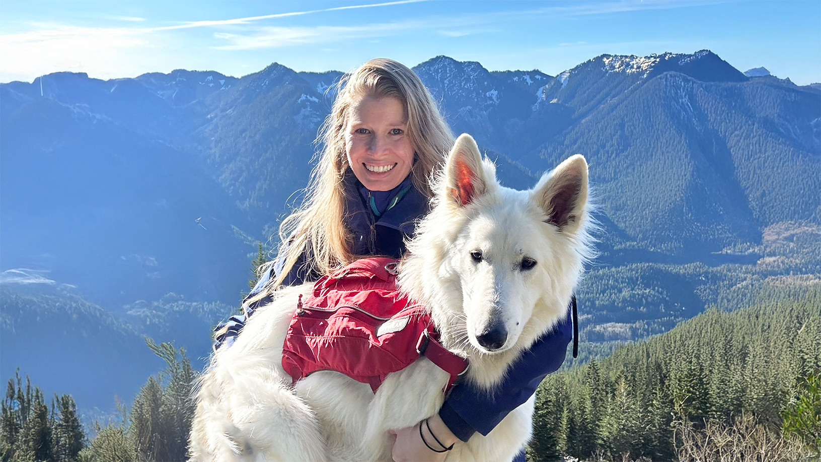 A woman holds a large white dog with mountains in the background.