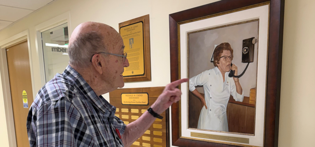 Dr. Cheney at the office with a photo of his mother, Laura hanging on the wall. She is in a nurses' uniform, talking on the phone.