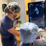Two healthcare professionals in scrubs and masks operate an ultrasound machine in a clinical setting, reviewing the monitor display.