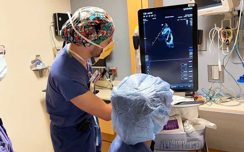 Two healthcare professionals in scrubs and masks operate an ultrasound machine in a clinical setting, reviewing the monitor display.