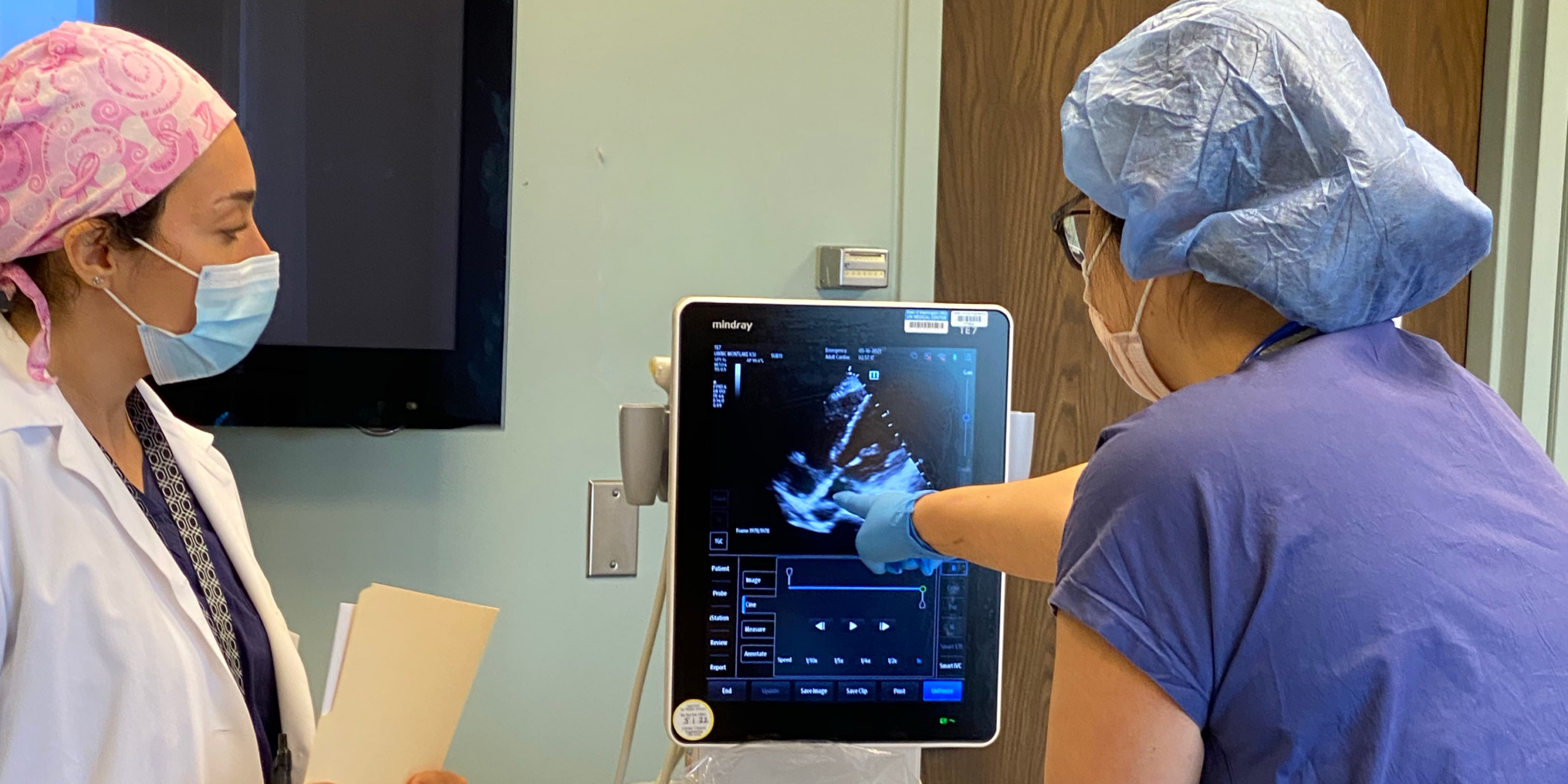 Two healthcare professionals in scrubs and masks operate an ultrasound machine in a clinical setting, reviewing the monitor display.